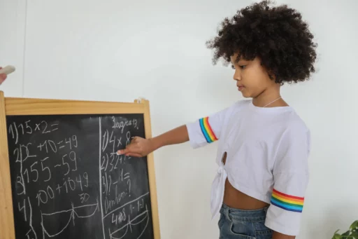 A young girl writing math problems on a small chalkboard. She is focused and engaged, wearing a white shirt with rainbow stripes on the sleeves.