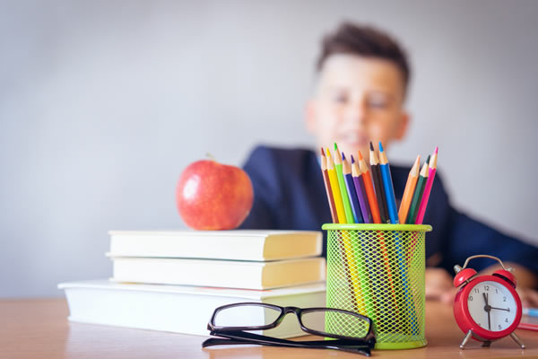 Colorful pencils in a holder with books, an apple, glasses, and an alarm clock on a desk, with a blurred student in the background.
