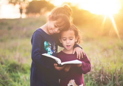 Two young girls reading a book together outdoors at sunset. One girl has her arm around the other, and they are both focused on the book, independent learning. This shows away to encourage independent learning