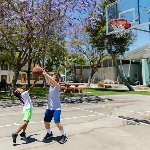 Two children playing basketball on an outdoor court. The child is attempting to shoot while the adult is blocking. The scene is set under trees with purple blossoms and clear blue skies in the background.